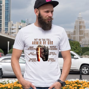May include: White t-shirt with a graphic of a knight in armor holding a sword. The text "Put on the full Armor of God" is printed on the shirt, along with the words "Helmet of Salvation", "Breastplate of Righteousness", "Belt of Truth", "Shield of Faith", "Sword of Spirit", and "Shoes of Peace".