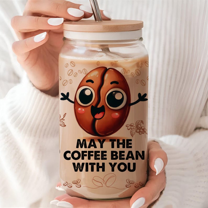 May include: A glass jar with a wooden lid filled with iced coffee. The jar has a cartoon coffee bean with a smiling face and the text "May the coffee bean with you".