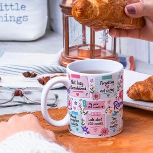 May include: White ceramic mug with a handle, decorated with colorful sticky notes and floral accents. The notes feature phrases like "Not lazy. Overwhelmed" and "Tiny starts matter." A croissant is held near the mug.