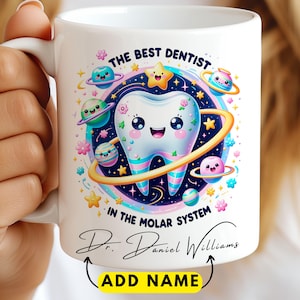 May include: White ceramic mug with a colorful cartoon design. The design features a smiling tooth surrounded by planets and stars, with the text "THE BEST DENTIST IN THE MOLAR SYSTEM" and a space for a name. The mug has a white handle.