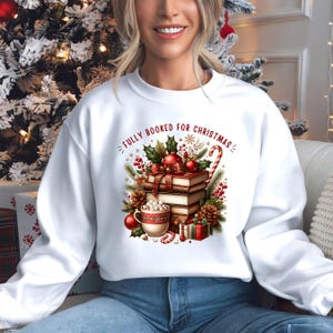 Op de afbeelding: Witte sweatshirt met een feestelijke afbeelding met de tekst "FULLY BOOKED FOR CHRISTMAS". Het ontwerp toont een stapel boeken, een mok marshmallows, ornamenten, dennenappels en een zuurstok. Het thema is Kerstmis en lezen.