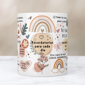 Op de afbeelding: Witte keramische mok versierd met pastelkleurige illustraties en Spaanse tekst. Het ontwerp bevat regenbogen, bloemen, harten en de zin "Recordatorios para cada día". Een motiverend en inspirerend cadeau.
