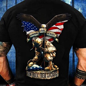 May include: Black t-shirt featuring a patriotic design with an eagle, American flag, and a kneeling soldier. The text reads "Honoring Our Heroes" and "Remember Their Sacrifice". The design is detailed and rendered in shades of brown, red, white, and blue.