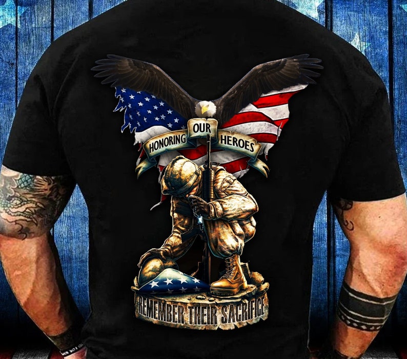 May include: Black t-shirt featuring a patriotic design with an eagle, American flag, and a kneeling soldier. The text reads "Honoring Our Heroes" and "Remember Their Sacrifice". The design is detailed and rendered in shades of brown, red, white, and blue.