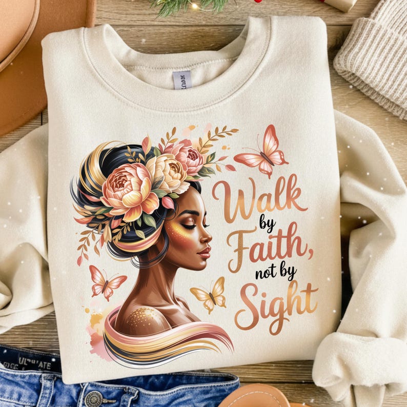 May include: Cream-colored sweatshirt featuring a graphic of a woman with floral headwear and butterflies. The text "Walk by Faith, not by Sight" is displayed in a stylish font. The design incorporates warm colors.