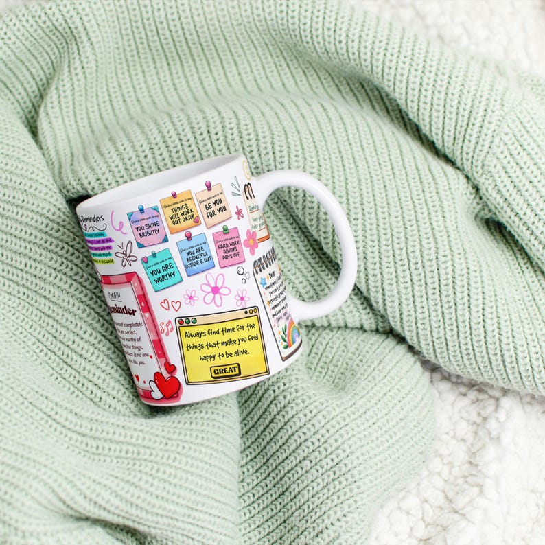 May include: White ceramic mug with colorful inspirational notes and a heart design. The mug features text like "You are worthy" and "Always find time for the things that make you feel happy to be alive." The mug is resting on a light green knitted blanket.