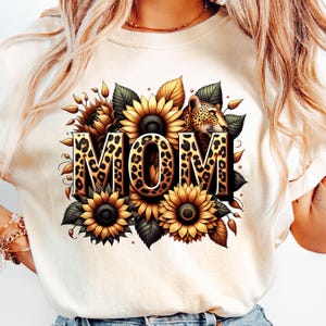 May include: White t-shirt with a graphic design of the word "MOM" in leopard print surrounded by sunflowers and a leopard head.