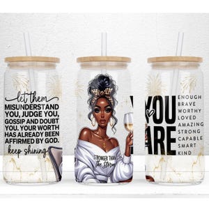 May include: Three clear glass tumblers with bamboo lids and straws. Each tumbler features a different design. The first tumbler has a black woman with a white shirt and the text "let them misunderstand you, judge you, gossip and doubt you. Your worth has already been affirmed by God. keep shining". The second tumbler has a black woman with a white shirt and the text "Stronger than the storm". The third tumbler has the text "You are" with the words "enough brave worthy loved amazing strong capable smart kind" listed below.