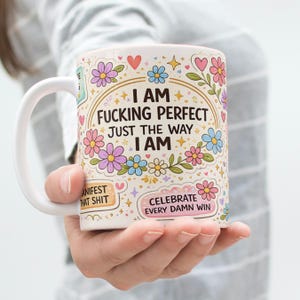 May include: White ceramic mug with a curved handle, featuring a colorful floral design and the text "I AM FUCKING PERFECT JUST THE WAY I AM". Additional text includes "MANIFEST THAT SHIT" and "CELEBRATE EVERY DAMN WIN".