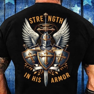 Puede incluir: Camiseta negra con un diseño de caballero. El gráfico muestra un caballero con armadura, alas, escudos y una espada. El texto dice "STRENGTH IN HIS ARMOR" en una fuente decorativa.