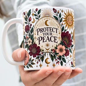 May include: A white ceramic mug featuring a central oval frame with the text "PROTECT YOUR PEACE" in black lettering. The design includes a golden sun, crescent moons, stars, and dark red and pink floral illustrations with green leaves.