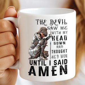May include: White ceramic mug featuring a kneeling knight illustration in silver armor, holding a sword. The mug has the text "THE DEVIL SAW ME WITH MY HEAD DOWN AND THOUGHT HE'D WON UNTIL I SAID AMEN" in black font.