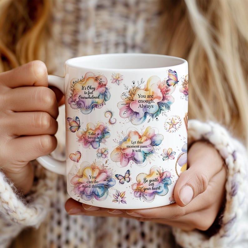 K&ouml;nnte beinhalten: Wei&szlig;er Keramikbecher mit bunten Blumendesigns, Schmetterlingen und inspirierenden Zitaten. Der Becher zeigt S&auml;tze wie "Du bist genug" und "Sei sanft zu deinem Herzen."