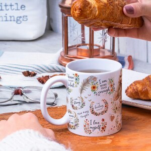 May include: White ceramic mug with a curved handle, decorated with floral designs and positive affirmations. The mug is filled with tea and sits on a wooden surface. A croissant is held nearby.