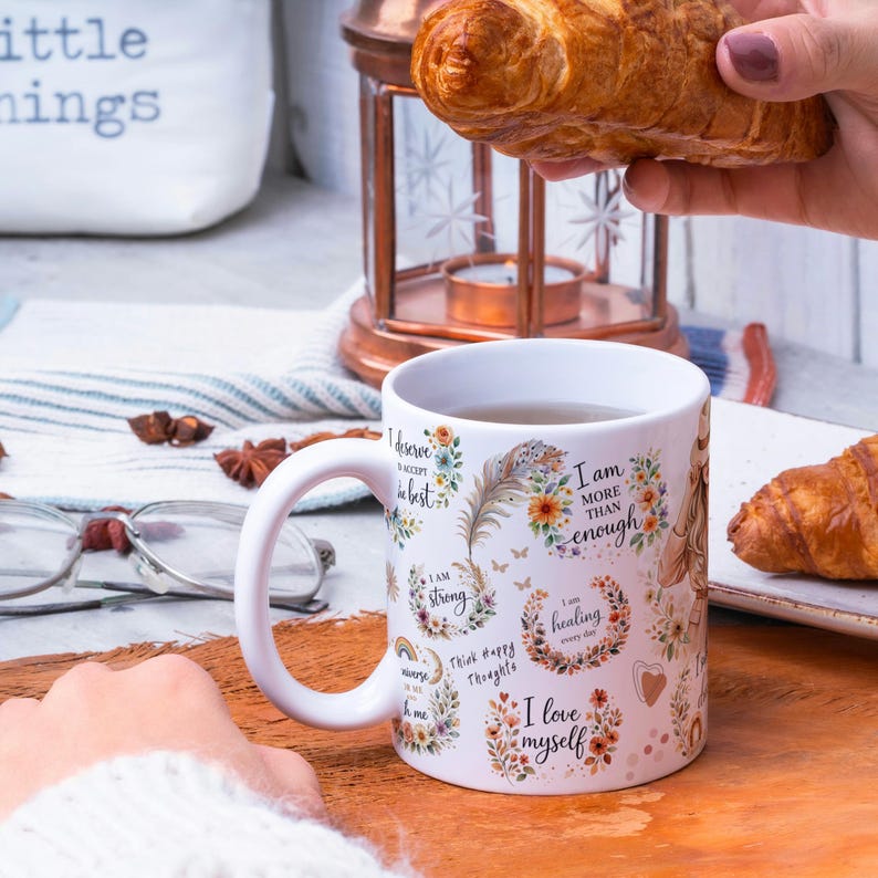 May include: White ceramic mug with a curved handle, decorated with floral designs and positive affirmations. The mug is filled with tea and sits on a wooden surface. A croissant is held nearby.