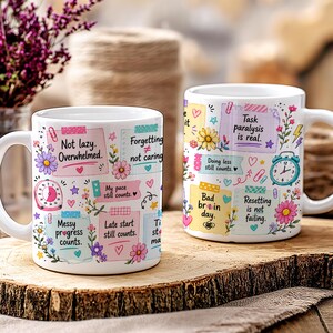 May include: Two white ceramic mugs with colorful designs and text. One mug features phrases like "Not lazy. Overwhelmed." and "Messy progress counts." The other mug has "Task paralysis is real." and "Bad brain day." Floral and heart accents.