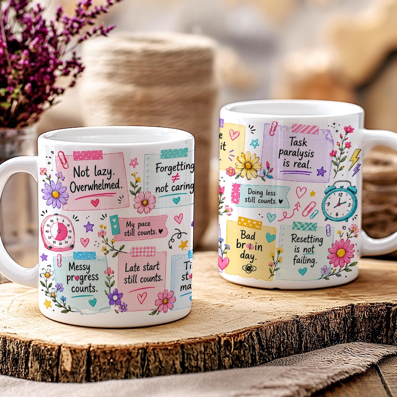 May include: Two white ceramic mugs with colorful designs and text. One mug features phrases like "Not lazy. Overwhelmed." and "Messy progress counts." The other mug has "Task paralysis is real." and "Bad brain day." Floral and heart accents.