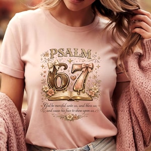 May include: A light pink t-shirt featuring the text "PSALM 67" in a decorative font, with floral and bow accents. The design includes an open book and the phrase "God be merciful unto us, and bless us; and cause his face to shine upon us."