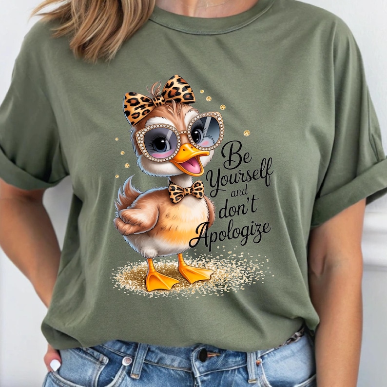 May include: Olive green t-shirt featuring a cartoon duck wearing sunglasses, a bow, and a bow tie. The duck has a brown body, yellow feet, and a yellow beak. Text on the shirt reads "Be Yourself and don't Apologize."