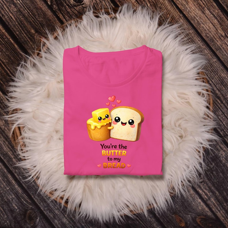 May include: A bright pink t-shirt featuring a cute cartoon illustration of a stick of butter and a slice of bread with smiling faces. The text "You're the Butter to my Bread" is printed below the characters, with small hearts.