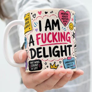 May include: A white ceramic mug featuring the text I AM A FUCKING DELIGHT in black and pink lettering. The design includes colourful doodles, hearts, stars, a yellow crown, a smiley face, and text boxes reading Mental Health Matters, Bitch and Tiny Wins Count A Lot.