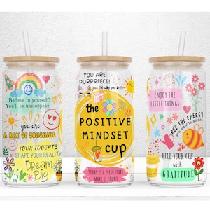 Puede incluir: Tres vasos de vidrio coloridos con citas inspiradoras e ilustraciones. Los vasos tienen tapas de bambú y pajitas. Las citas son "¡Cree en ti mismo! ¡Serás imparable! Eres un rayo de sol. Tus pensamientos dan forma a tu realidad. Sueña a lo grande", "¡Eres perfecto! Tal como eres! La taza de mentalidad positiva. Hoy es un nuevo comienzo. Haz que cuente", y "Disfruta de las pequeñas cosas. Sé la energía que quieres atraer. Llena tu taza de gratitud".
