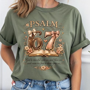 May include: Olive green t-shirt with the text "PSALM 67" in a decorative font. The design includes the numbers 6 and 7, adorned with bows and floral accents, and an open book. The shirt also features a quote: "God be merciful unto us, and bless us; and cause his face to shine upon us."