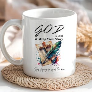 Puede incluir: Taza de cer&aacute;mica blanca con un dise&ntilde;o colorido que presenta una pluma estilogr&aacute;fica, una cruz y un rollo. El texto dice "GOD is still Writing Your Story Stop trying to steal the pen."