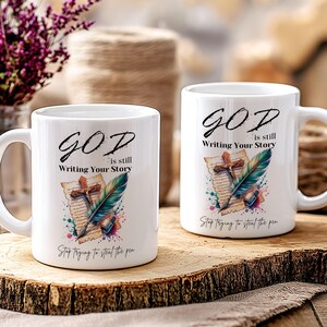 Puede incluir: Dos tazas de cer&aacute;mica blanca con un dise&ntilde;o colorido que presenta una pluma estilogr&aacute;fica, una cruz y un trozo de pergamino. El texto en las tazas dice "GOD is still Writing Your Story" y "Stop trying to steal the pen".