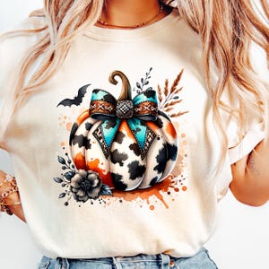 May include: Cream-colored t-shirt featuring a watercolor pumpkin design. The pumpkin has a cow print pattern, an orange and turquoise bow, and is decorated with floral and bat accents. A fall-themed graphic tee.