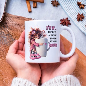 K&ouml;nnte beinhalten: Wei&szlig;e Keramik-Kaffeetasse mit einer Cartoon-Illustration eines M&auml;dchens, das eine gro&szlig;e Kaffeetasse umarmt. Auf der Tasse steht in Rosa und Schwarz der Text "I'M UP... THE REST OF THE DAY BROUGHT TO YOU BY COFFEE!!".