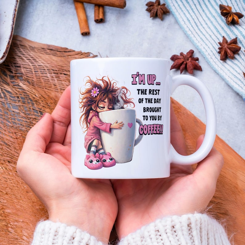 K&ouml;nnte beinhalten: Wei&szlig;e Keramik-Kaffeetasse mit einer Cartoon-Illustration eines M&auml;dchens, das eine gro&szlig;e Kaffeetasse umarmt. Auf der Tasse steht in Rosa und Schwarz der Text "I'M UP... THE REST OF THE DAY BROUGHT TO YOU BY COFFEE!!".