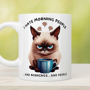 Puede incluir: Taza de cer&aacute;mica blanca con una ilustraci&oacute;n de un gato gru&ntilde;&oacute;n y el texto "I hate morning people...and mornings...and people." El gato tiene una expresi&oacute;n enfadada y sostiene una taza de caf&eacute; azul.