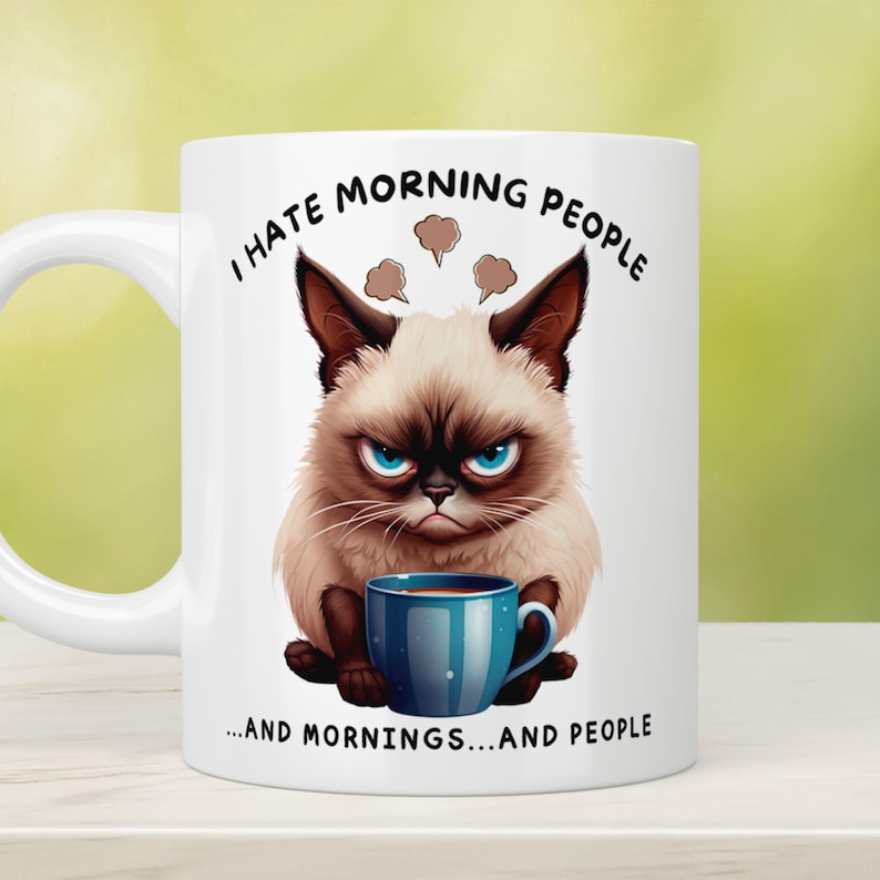 Puede incluir: Taza de cer&aacute;mica blanca con una ilustraci&oacute;n de un gato gru&ntilde;&oacute;n y el texto "I hate morning people...and mornings...and people." El gato tiene una expresi&oacute;n enfadada y sostiene una taza de caf&eacute; azul.