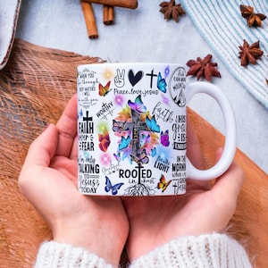 May include: White ceramic mug with a cross, butterflies, and inspirational text. The design includes phrases like "Faith over Fear" and "Rooted in Christ." The mug has a white handle and is held by a person.