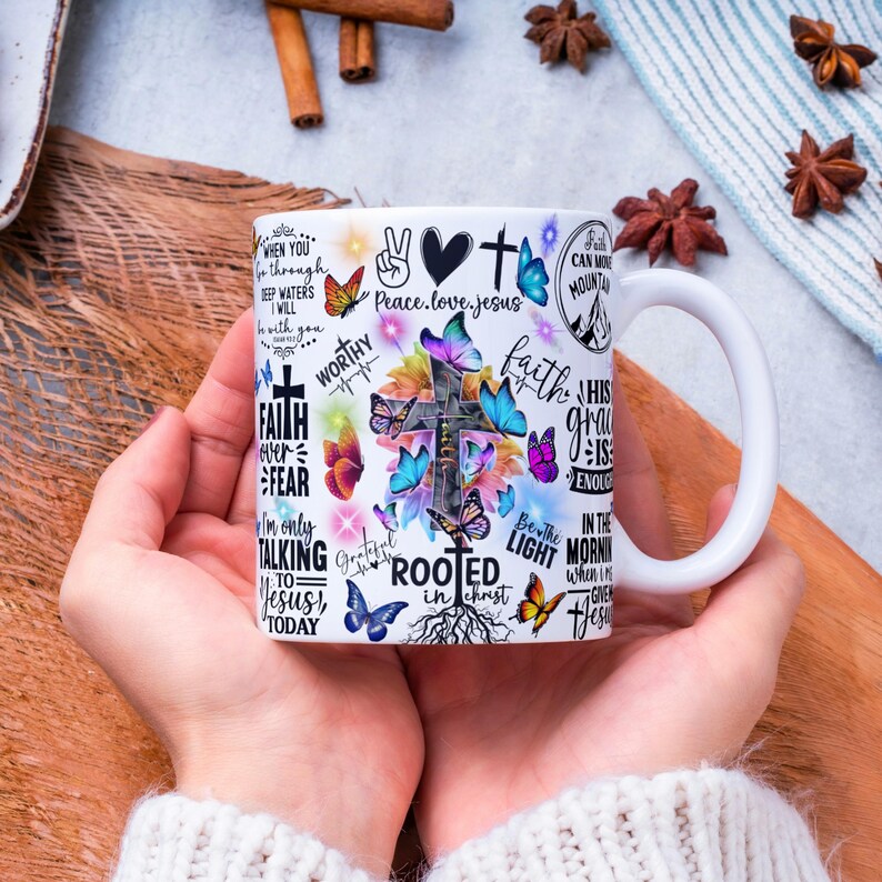 May include: White ceramic mug with a cross, butterflies, and inspirational text. The design includes phrases like "Faith over Fear" and "Rooted in Christ." The mug has a white handle and is held by a person.