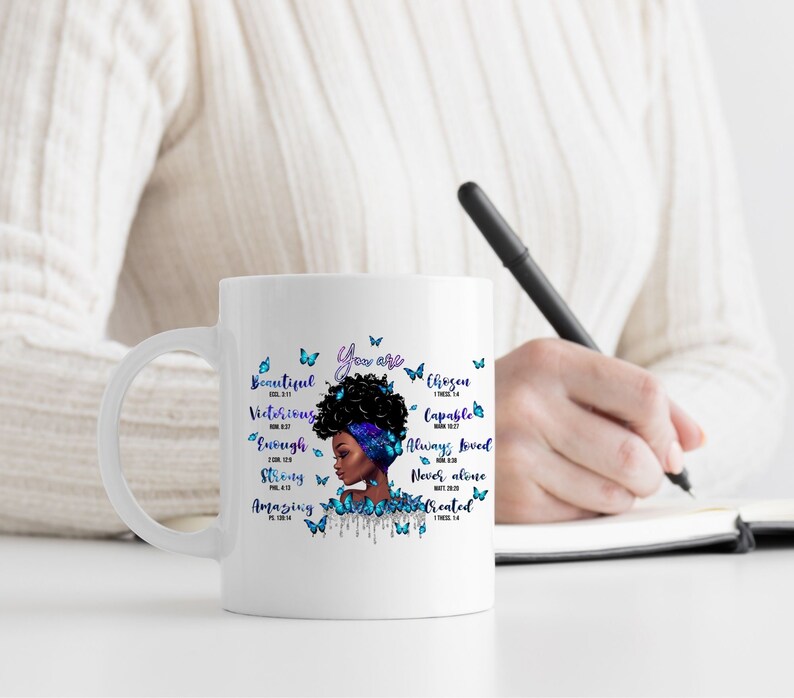 May include: White ceramic mug with a black and blue illustration of a woman with an afro and butterflies. The mug has the text "You are" and several words with bible verses, including "Beautiful", "Victorious", "Chosen", "Capable", "Enough", "Always Loved", "Never Alone", "Strong", "Amazing", and "Created".