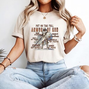 May include: A beige t-shirt featuring a graphic of a knight in armour and the text "Put on the full Armor of God". The graphic includes the text "Helmet of Salvation", "Breastplate of Righteousness", "Belt of Truth", "Shield of Faith", "Sword of Spirit", and "Shoes of Peace".