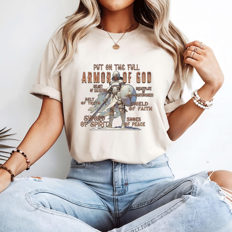 May include: A beige t-shirt featuring a graphic of a knight in armour and the text "Put on the full Armor of God". The graphic includes the text "Helmet of Salvation", "Breastplate of Righteousness", "Belt of Truth", "Shield of Faith", "Sword of Spirit", and "Shoes of Peace".