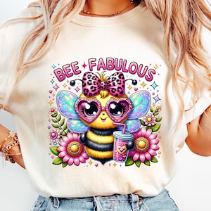 May include: A cream-colored t-shirt featuring a cartoon bee with heart-shaped sunglasses, a bow, and wings. The bee is holding a drink and surrounded by flowers. The text "BEE FABULOUS" is above the bee.