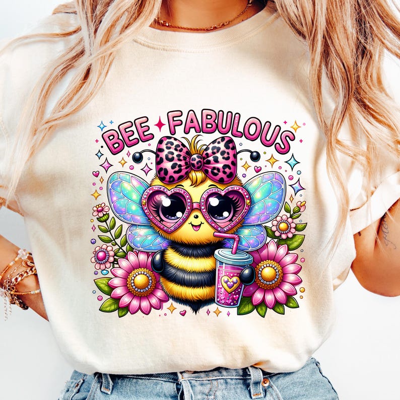May include: A cream-colored t-shirt featuring a cartoon bee with heart-shaped sunglasses, a bow, and wings. The bee is holding a drink and surrounded by flowers. The text "BEE FABULOUS" is above the bee.
