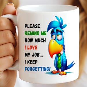 May include: White ceramic mug with the text "PLEASE REMIND ME HOW MUCH I LOVE MY JOB... I KEEP FORGETTING!" and a cartoon blue parrot with a yellow beak and green tie. The mug is held by a person.
