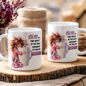 K&ouml;nnte beinhalten: Zwei wei&szlig;e Keramik-Kaffeetassen mit dem Text "I'M UP... THE REST OF THE DAY BROUGHT TO YOU BY COFFEE!!" und einer Cartoon-Illustration eines M&auml;dchens, das eine Kaffeetasse umarmt. Das M&auml;dchen tr&auml;gt einen rosa Kapuzenpullover und rosa Hausschuhe.