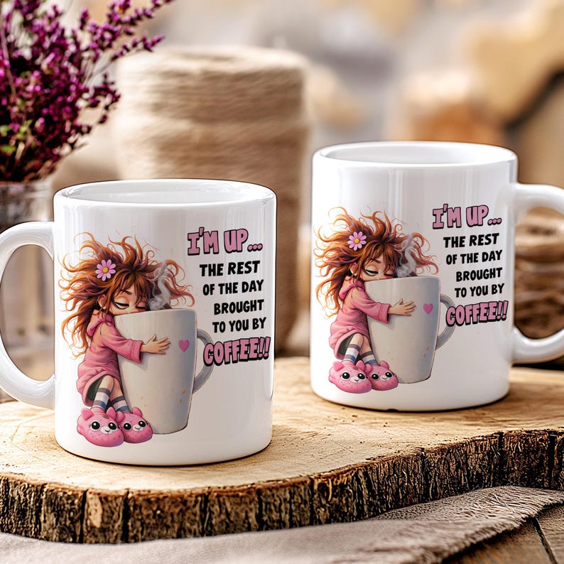 K&ouml;nnte beinhalten: Zwei wei&szlig;e Keramik-Kaffeetassen mit dem Text "I'M UP... THE REST OF THE DAY BROUGHT TO YOU BY COFFEE!!" und einer Cartoon-Illustration eines M&auml;dchens, das eine Kaffeetasse umarmt. Das M&auml;dchen tr&auml;gt einen rosa Kapuzenpullover und rosa Hausschuhe.