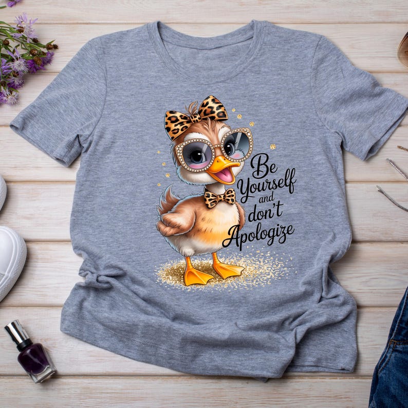 May include: A light gray t-shirt featuring a cartoon duck wearing sunglasses, a bow, and a bow tie. The duck is standing on a patch of gold glitter. Text on the shirt reads "Be Yourself and don't Apologize."