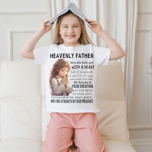 May include: White t-shirt with a graphic of a girl praying and the text "HEAVENLY FATHER bless this little girl WITH A HEART full of kindness. a spirit of joy AND EYES THAT SEE the beauty in YOUR CREATION MAY SHE GROW IN YOUR LOVE, surrounded by your grace, AND FIND STRENGTH IN YOUR PRESENCE"