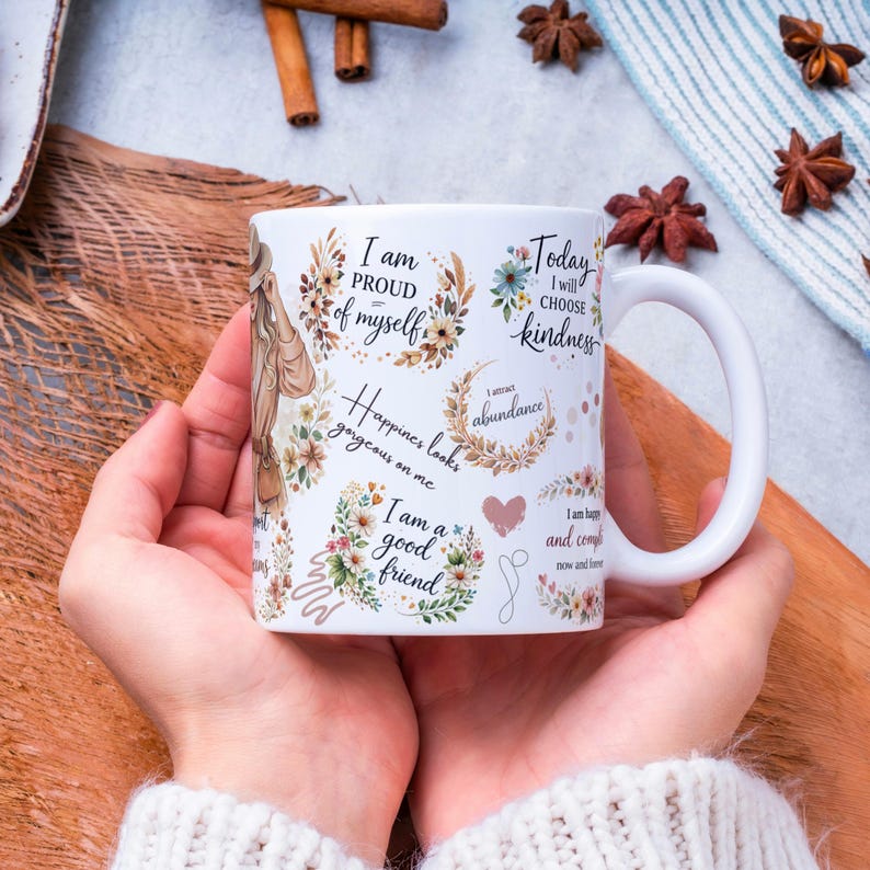 May include: White ceramic mug with a curved handle, decorated with floral wreaths and inspirational phrases like "I am proud of myself" and "Today I will choose kindness." The mug is held in two hands.