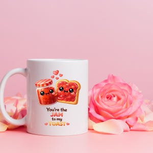 May include: White ceramic mug with a handle, featuring a cartoon illustration of a jam jar and a slice of toast with smiling faces and hearts. The text "You're the JAM to my TOAST" is printed below the images. A pink rose is in the background.