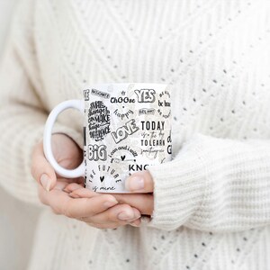 May include: White ceramic mug with black handwritten inspirational words like "love", "today", and "choose". The mug is held by a person wearing a white sweater.
