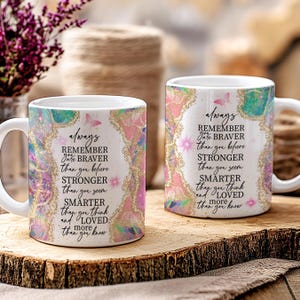 Op de afbeelding: Twee witte keramische mokken met de tekst "Onthoud altijd dat je moediger bent dan je gelooft, sterker dan je lijkt, slimmer dan je denkt en meer geliefd dan je weet." De mokken hebben een kleurrijk bloemmotief.